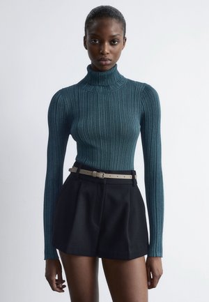 Woman wearing a teal ribbed turtleneck sweater paired with high-waisted black shorts and a beige belt, standing against a plain white wall.