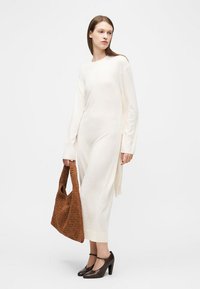 Femme portant une robe blanche à manches longues et longueur cheville, tenant un grand sac marron texturé, chaussée de chaussures à talons hauts marron foncé.