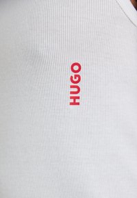 Débardeur blanc à côtes présentant un logo rouge "HUGO" bien en évidence. La texture du tissu est fine avec des lignes verticales, créant une apparence ajustée.