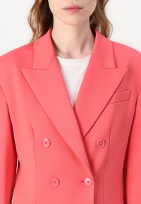 Koraal dubbelzijdige blazer met een scherpe revers, textuurstof en twee bijpassende knopen. Beschikt over een klein borstzakje.