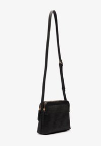 Schwarze Umhängetasche mit geflochtenem Textur, Reißverschluss und verstellbarem Trageriemen. verfügt über goldfarbene Hardware und rechteckige Form.