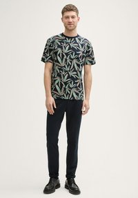 Zwarte T-shirt met korte mouwen met een bladpatroon in groen en roze, gecombineerd met donkere broeken en zwarte schoenen.