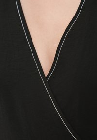 Μαύρο τοπ τύπου wrap με βαθύ V λαιμόκοψη, με αντίθετη λευκή ραφή κατά μήκος των άκρων, κατασκευασμένο από ελαφρύ ύφασμα.