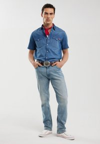 Camicia in denim di colore blu medio con maniche corte, due tasche sul petto, abbinata a jeans blu chiaro e sneakers bianche con dettagli rossi.