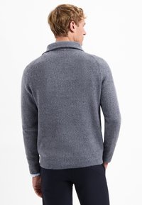 Blonde man die wegkijkt, in een gestructureerde grijze gebreide trui met een sjalkraag en donkere broeken tegen een witte achtergrond.