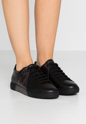 Zwarte lage leren sneakers met gestructureerde zijpanelen en zwarte veters, gedragen op blote voeten tegen een effen achtergrond.