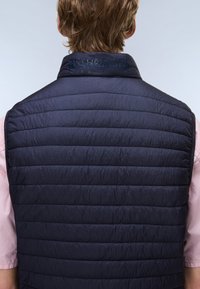 Mladý človek s krátkymi vlasmi, ktorý má na sebe tmavomodrú prešívanú bezrukávovú vestu cez svetloružovú košeľu, zobrazený zozadu.