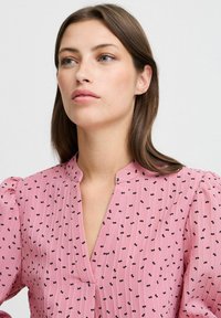 Rosa Bluse mit kleinen schwarzen Schleifenmustern, einem Kragen und gepufften Ärmeln. Strukturierter Stoff mit einer Raffung vorne.