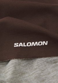 Brun stoff med en jevn tekstur, som har en hvit "SALOMON" logo. Kanten overgår til et lysegrått stoff nedenfor.