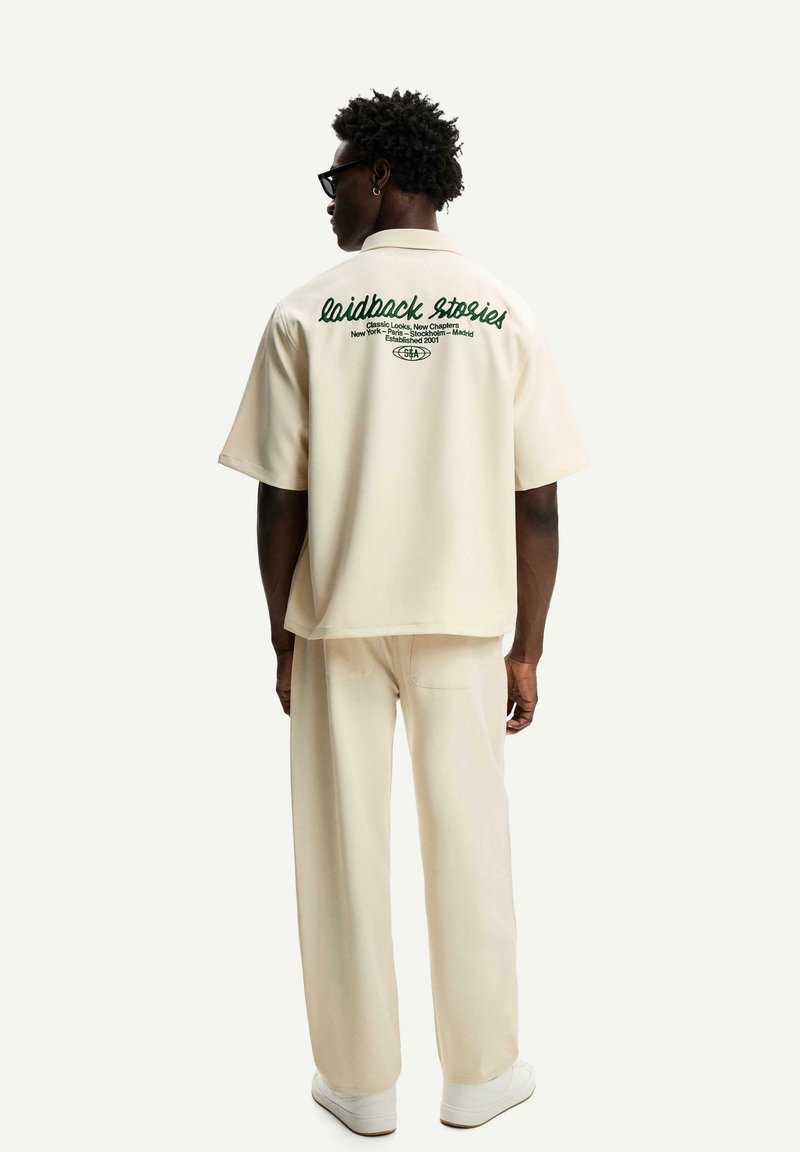 Uomo con capelli scuri e ricci e occhiali da sole, che indossa una camicia breve crema e pantaloni con testo verde sulla schiena, su uno sfondo bianco.