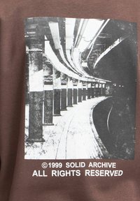 Brun sweatshirt med et sort-hvidt fotoprint af en metrostation med buede skinner og betonpiller. Teksten nederst lyder ©1999 SOLID ARCHIVE ALL RIGHTS RESERVED.
