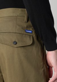 Pantalons vert olive avec une texture lisse, présentant une poche arrière, une fermeture à bouton, et un patch logo « HUGO » bleu.
