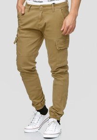 Khaki cargo hlače s elastičnim manžetama, višestrukim džepovima i zatvaranjem na patentni zatvarač. Uparene su s bijelim tenisicama i vidljivim Calvin Klein pojasom.