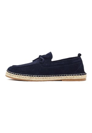 Zapato azul marino de ante tipo mocasín con suela envuelta en yute y detalle decorativo de nudo sobre fondo blanco.