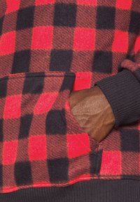 Rot-schwarz karierte Fleece-Hoodie mit Kängurutasche und schwarz gerippten Bündchen, zeigt eine weiche Textur und auffälliges Muster.