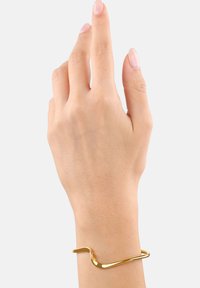 Pulsera de metal dorado con un diseño ondulado, textura suave y un extremo abierto, llevada en una mano con esmalte de uñas natural.