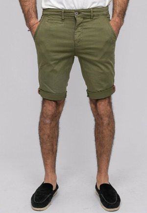 Homme portant un short vert olive retroussé et des chaussures noires sans lacets, debout les mains dans les poches devant un fond uni.
