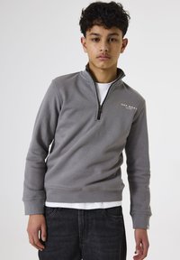 Joven adolescente con cabello rizado, vistiendo un suéter gris de medio cierre sobre una camiseta blanca y jeans negros, de pie contra un fondo liso.