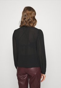 Blouse noire à manches longues avec un motif à carreaux texturé, dos transparent et manches froncées. Portée sur un pantalon en similicuir bordeaux.