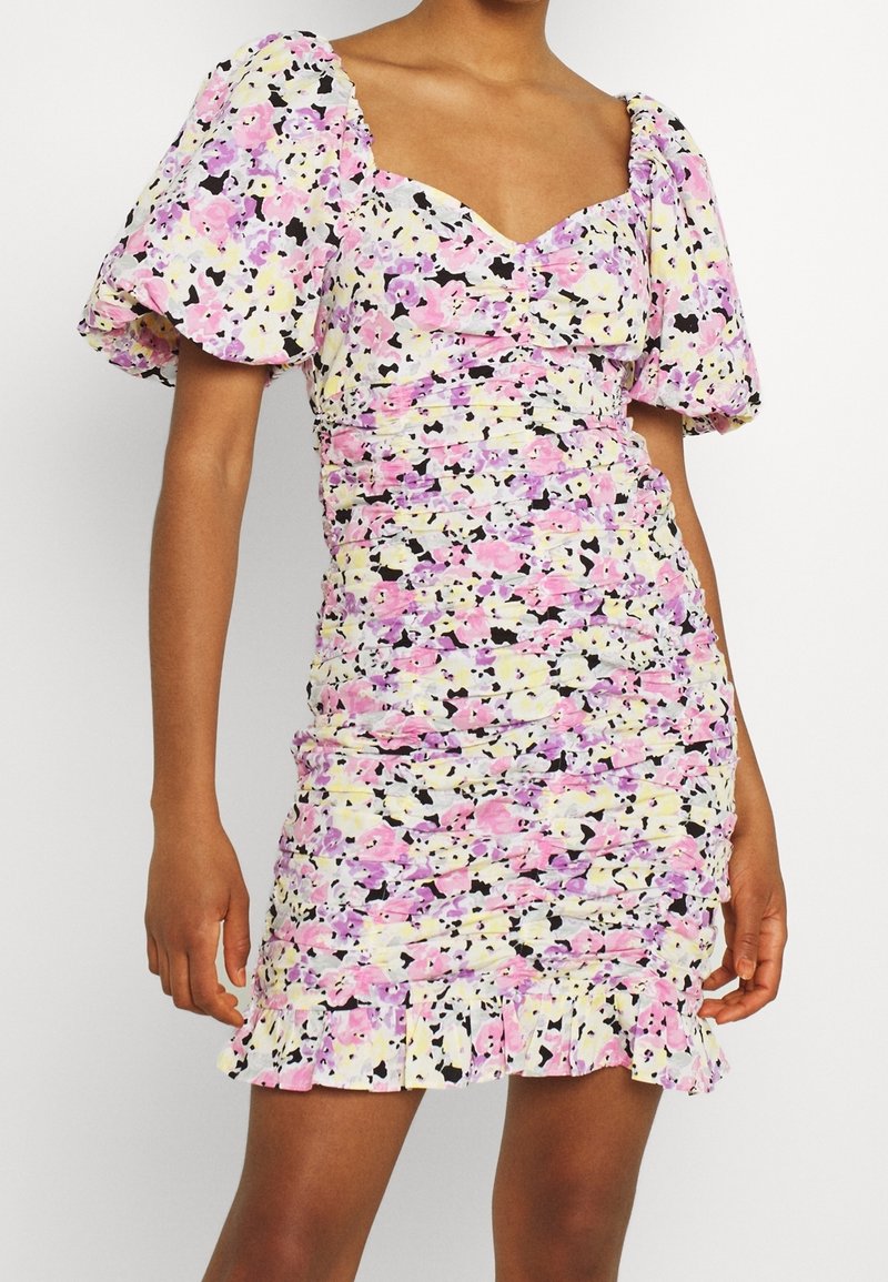 Šaty s květinovým potiskem, nafouklými rukávy, nařaseným designem a volánkovým lemem. Barvy zahrnují růžovou, žlutou a černou na bílém pozadí.