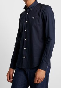 Laivastonsininen pitkähihaisen paita nappikauluksella, jossa pienen valkoisen logon rinnassa ja vaaleat napit edessä napituksessa.