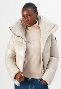 Beige pufferjack met hoge kraag, glanzende afwerking en horizontale stiksels. Draagt over een lichtbeige turtleneck en blauwe jeans.
