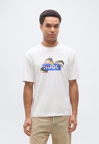 Άνδρας που φοράει λευκό μπλουζάκι με πολύχρωμα ψάρια και μπλε λογότυπο «HUGO», σε συνδυασμό με μπεζ παντελόνι, στέκεται μπροστά σε απλό ανοιχτό γκρι φόντο.