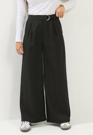 Kobieta ubrana w czarne, szerokie spodnie z wysokim stanem i paskiem z podwójnym pierścieniem, biały top z długim rękawem oraz białe trampki z czarnymi paskami.