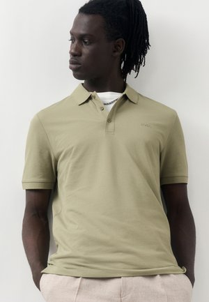 Άνδρας με dreadlocks κοιτάζει προς τα πλάγια φορώντας ανοιχτοπράσινο πόλο πάνω από λευκό μπλουζάκι και μπεζ παντελόνι, στέκεται δίπλα σε λευκό τοίχο.