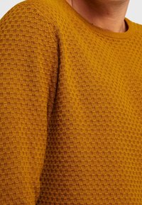 Pull moutarde avec un motif alvéolé texturé, col côtelé et manches longues. Le tissu semble doux et chaud.