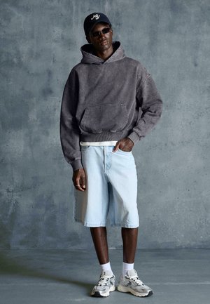 Moški, oblečen v temno sivo kapuco, svetlo modre kratke hlače do kolen, bele superge, črna sončna očala in mornarsko modro kapo na sivi teksturirani podlagi.