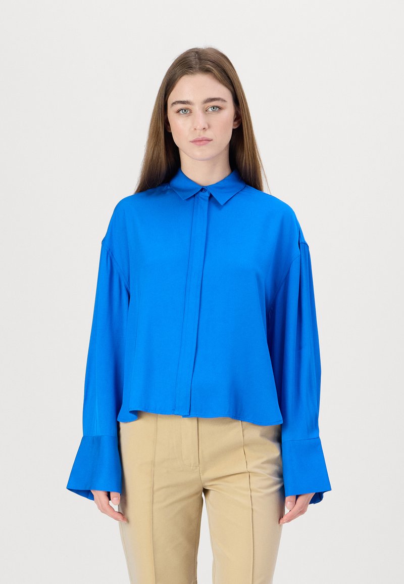Blue collared button-up shirt with long, loose sleeves and flared cuffs, made of smooth fabric; paired with beige trousers.
