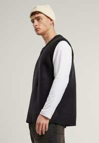 Dark grey sleeveless sweater with a ribbed neckline, worn over a white long-sleeve shirt. Cotton material, relaxed fit, minimal design.