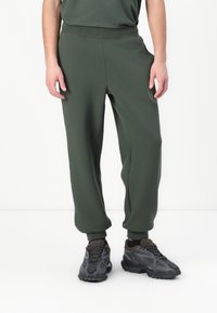 Pantalones deportivos de color verde oscuro con un ajuste clásico, cintura elástica y puños acanalados. Presenta un sutil parche con el logo en el muslo izquierdo.
