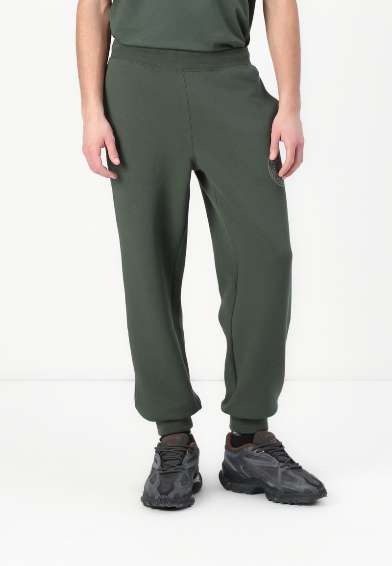 Pantalones deportivos de color verde oscuro con un ajuste clásico, cintura elástica y puños acanalados. Presenta un sutil parche con el logo en el muslo izquierdo.
