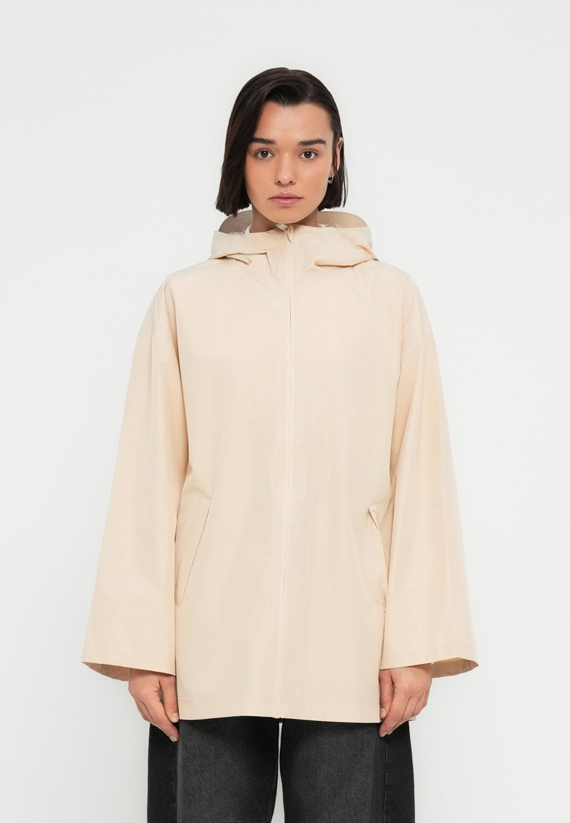 Imperméable beige clair avec capuche, doté d'une fermeture éclair, de deux poches latérales et d'une coupe décontractée. Texture lisse et design minimaliste.