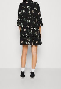 Schwarzes Kleid mit Blumenmustern, gestuftem Design, Dreiviertelärmeln und Rüschensaum. Getragen mit schwarzen Sneakern und weißen Socken.