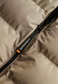 Beige gewatteerde jas met een zwarte ritssluiting en een opvallend oranje-geaccentueerd logo label, dat een gladde, glanzende textuur laat zien.