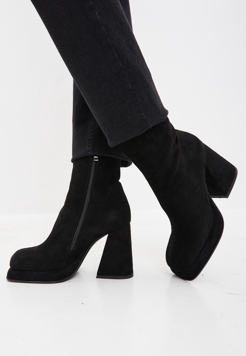 Black suede ankle boots with a block heel, featuring a side zipper and a rounded toe, paired with dark jeans on a white background.