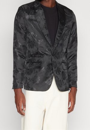 Zwart vest met bloemenprint van satijnstof, met een enkele knoopsluiting, gekartelde revers en twee voorzakken. Gekozen boven een zwart shirt.
