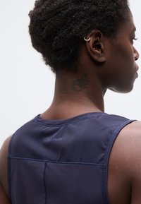 Κοντινό πλάνο του λαιμού και του άνω μέρους της πλάτης ενός ατόμου με ένα σκοτεινό τατουάζ, φορώντας μπλε αμάνικη μπλούζα και χρυσά σκουλαρίκια περιμετρικά του αυτιού.