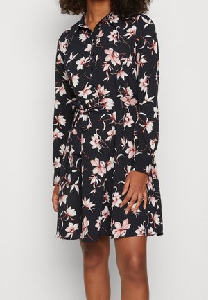 Frau trägt ein langärmliges, schwarzes Kleid mit Knöpfen, pink-weißen Blumenmustern und einem gebundenen Taillengürtel.