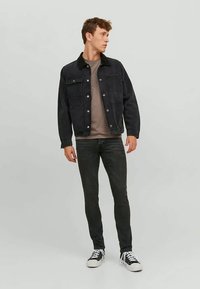 Young man in black denim jacket, grey shirt, black jeans, and black-and-white sneakers standing and looking right.