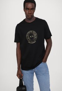T-shirt noir à manches courtes avec un logo rond en métal doré portant "A|X" et "ARMANI EXCHANGE." Le modèle porte un jean bleu clair et tient un sac.