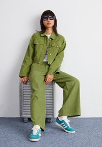 Groene corduroyjas en wijde broek, gecombineerd met een wit shirt en teal sneakers. Model draagt donkere zonnebril en zit op een grijs oppervlak.