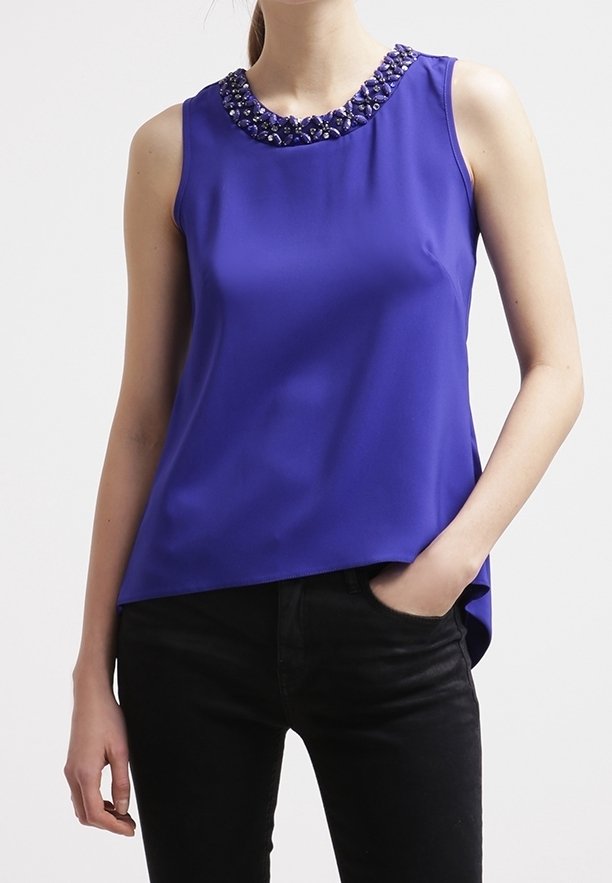 Haut sans manches bleu avec un col en perles, fabriqué en tissu lisse, présentant une coupe décontractée et un ourlet asymétrique. Porte avec un pantalon noir.