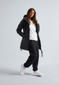 Zwarte pufferjas met een capuchon, gestikte ontwerp en een ritssluiting. Gecombineerd met een wit T-shirt, zwarte joggers en witte sneakers.