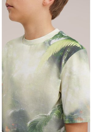 Dziecko w jasnooliwkowej koszulce z nadrukiem tropikalnych liści i motyla, zwrócone w lewo na neutralnym tle.