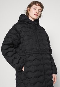 Zwarte gewatteerde jas met een kap. Voorzien van een quilted ontwerp, ritssluiting en zijzakken. Gemaakt van een gladde, waterafstotende stof.