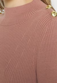 Pull en maille rose avec col montant, texture côtelée et trois boutons dorés en accent sur l'épaule gauche.